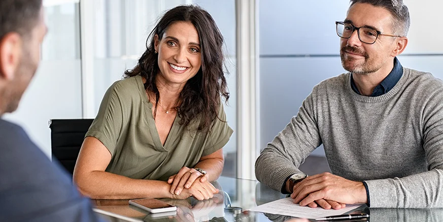 man and woman sit at a glass table with a phone and papers on it, in a meeting